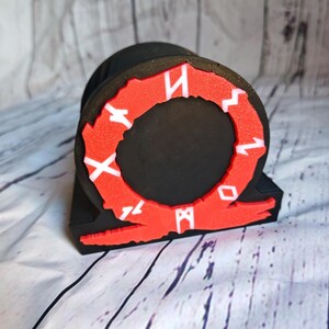 Peut inclure: Objet imprim&eacute; en 3D noir et rouge en forme de cercle avec des runes autour du bord. Les runes sont blanches et le cercle est rouge. L'objet est sur une surface en bois blanc.