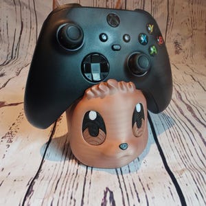 Peut inclure: Un support de manette imprim&eacute; en 3D de couleur marron et noir en forme d'animal de dessin anim&eacute; avec de grands yeux. Une manette Xbox noire repose sur le support.