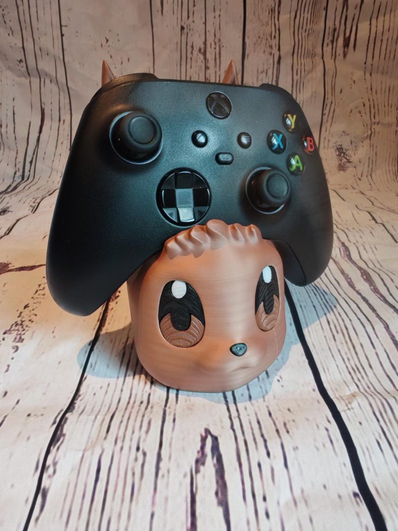 Peut inclure: Un support de manette imprim&eacute; en 3D de couleur marron et noir en forme d'animal de dessin anim&eacute; avec de grands yeux. Une manette Xbox noire repose sur le support.