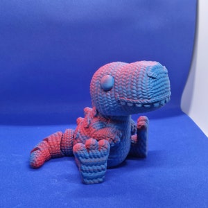 May include: A blue and pink 3D printed dinosaur toy. The dinosaur is sitting with its head tilted to the side and its mouth slightly open. The toy has a textured surface that resembles knitted fabric.