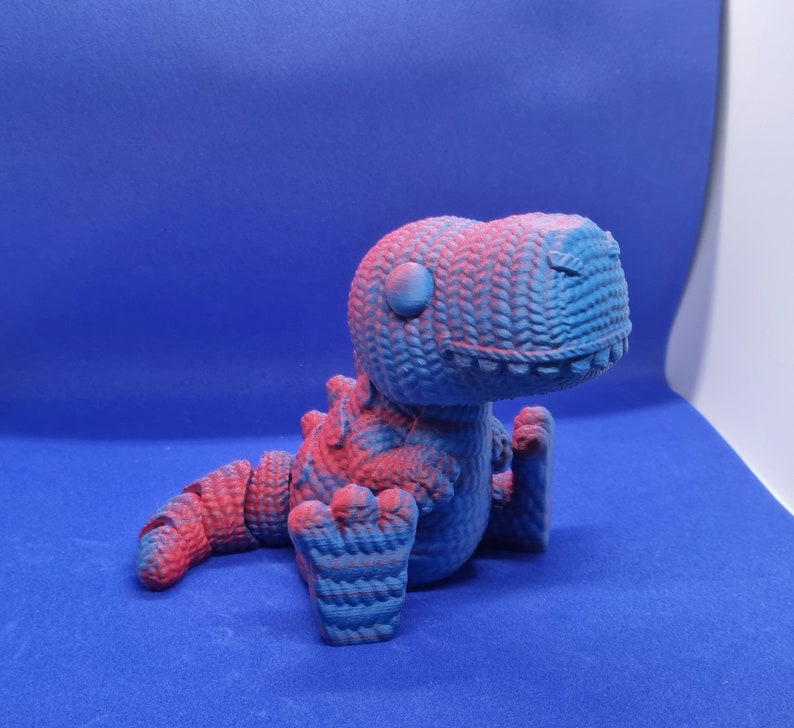 May include: A blue and pink 3D printed dinosaur toy. The dinosaur is sitting with its head tilted to the side and its mouth slightly open. The toy has a textured surface that resembles knitted fabric.
