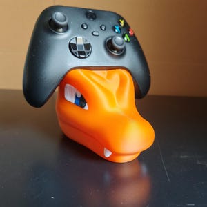 Op de afbeelding: Een zwarte Xbox-controller rust op een oranje, 3D-geprinte standaard in de vorm van een Pok&eacute;mon-personage. De standaard heeft een blauw oog en witte tanden.
