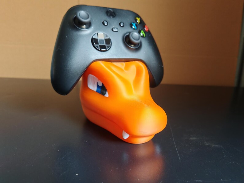 Op de afbeelding: Een zwarte Xbox-controller rust op een oranje, 3D-geprinte standaard in de vorm van een Pok&eacute;mon-personage. De standaard heeft een blauw oog en witte tanden.