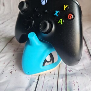 Op de afbeelding: Een zwarte Xbox-controller rust op een lichtblauwe en witte gameconsole-standaard. De controller heeft gekleurde knoppen en een wit Xbox-logo. De standaard heeft een gestileerd ontwerp.