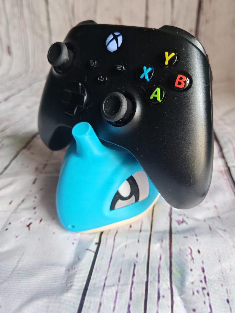 Op de afbeelding: Een zwarte Xbox-controller rust op een lichtblauwe en witte gameconsole-standaard. De controller heeft gekleurde knoppen en een wit Xbox-logo. De standaard heeft een gestileerd ontwerp.