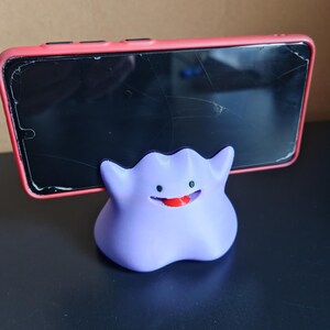 以下が含まれることがあります： ポケモンのキャラクターの形をした、紫と赤の3Dプリントされた携帯電話スタンド。スタンドは、ひび割れた画面の赤い電話を保持しています。