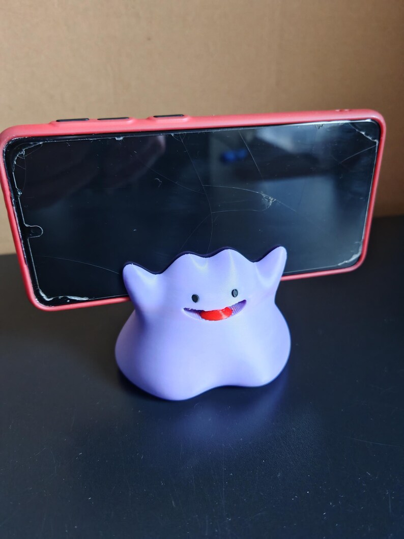 以下が含まれることがあります： ポケモンのキャラクターの形をした、紫と赤の3Dプリントされた携帯電話スタンド。スタンドは、ひび割れた画面の赤い電話を保持しています。