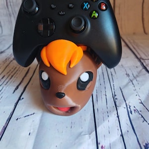 Könnte beinhalten: Eine braune und orangefarbene Pokemon-Figur mit einem schwarzen Xbox-Controller auf dem Kopf. Der Controller hat die Standard-Xbox-Tasten: A, B, X, Y.