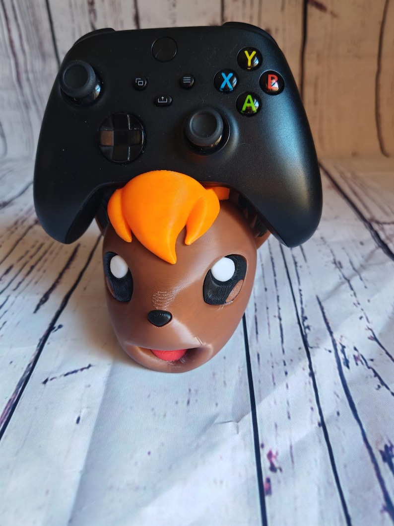 Pode incluir: Uma figura de personagem Pokemon marrom e laranja com um controlador Xbox preto no topo de sua cabe&ccedil;a. O controlador tem os bot&otilde;es Xbox padr&atilde;o: A, B, X, Y.