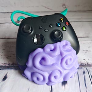 Op de afbeelding: Een zwarte Xbox-controller met een paarse en witte stander in de vorm van een wolk. De controller heeft een turquoise haak aan de bovenkant.