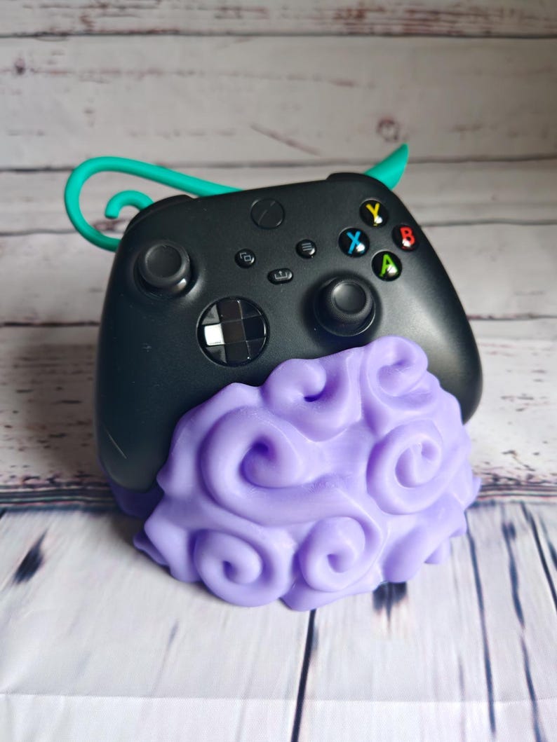 Op de afbeelding: Een zwarte Xbox-controller met een paarse en witte stander in de vorm van een wolk. De controller heeft een turquoise haak aan de bovenkant.