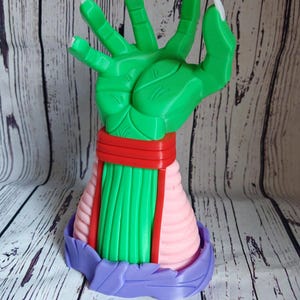 Op de afbeelding: Een groene hand met lange witte klauwen, met een rood-roze gestreept armband. De hand is in een grijpende beweging geposeerd. De hand is bevestigd aan een paarse basis.