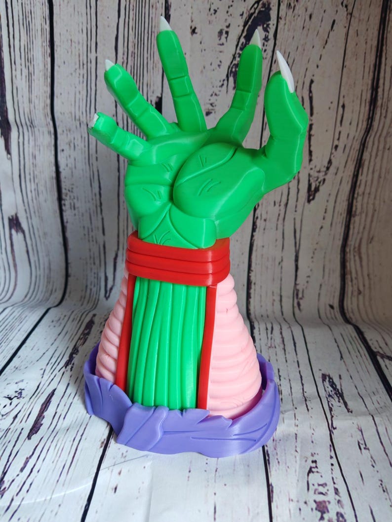 Op de afbeelding: Een groene hand met lange witte klauwen, met een rood-roze gestreept armband. De hand is in een grijpende beweging geposeerd. De hand is bevestigd aan een paarse basis.