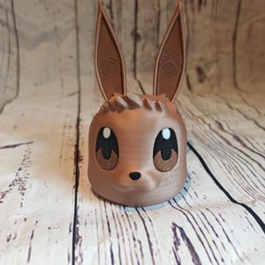 Peut inclure: Un pot en c&eacute;ramique brun en forme de lapin de dessin anim&eacute; avec de grands yeux et un petit nez. Le pot a une surface textur&eacute;e qui ressemble au bois.
