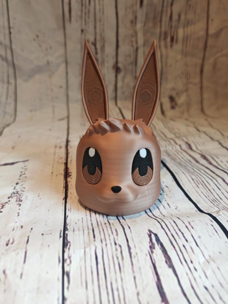 Peut inclure: Un pot en c&eacute;ramique brun en forme de lapin de dessin anim&eacute; avec de grands yeux et un petit nez. Le pot a une surface textur&eacute;e qui ressemble au bois.