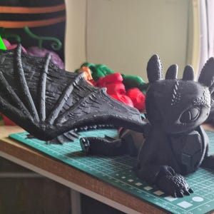 May include: A black 3D printed dragon figurine with large wings. The dragon has a textured surface and a detailed face.