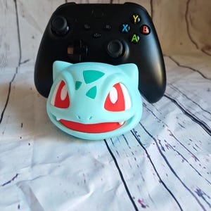 Op de afbeelding: Een zwarte videogamecontroller met kleurrijke knoppen zit bovenop een lichtblauw en rood Pokemon-figuur.