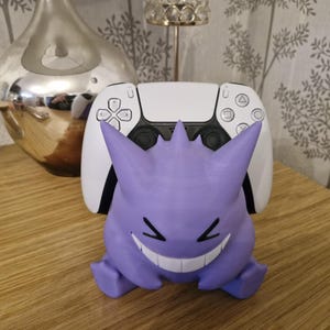 May include: A purple and white 3D printed Ghost Pokemon character holding a white Playstation 5 controller.