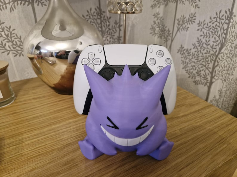 May include: A purple and white 3D printed Ghost Pokemon character holding a white Playstation 5 controller.
