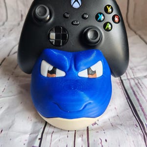 K&ouml;nnte beinhalten: Ein blau-wei&szlig;er, 3D-gedruckter Controller-Halter in Form eines Cartoon-Gesichts, mit einem schwarzen Xbox-Controller, der darauf ruht. Der Controller-Halter hat eine runde Basis und detaillierte Gesichtsz&uuml;ge.