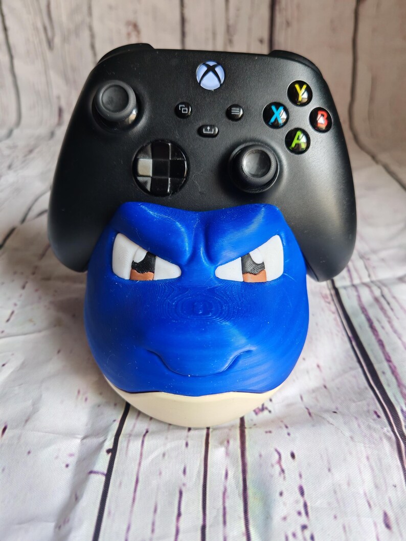 K&ouml;nnte beinhalten: Ein blau-wei&szlig;er, 3D-gedruckter Controller-Halter in Form eines Cartoon-Gesichts, mit einem schwarzen Xbox-Controller, der darauf ruht. Der Controller-Halter hat eine runde Basis und detaillierte Gesichtsz&uuml;ge.