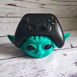 Op de afbeelding: Een zwarte Xbox-controller wordt in een groene Yoda-standaard gehouden. De controller heeft de standaard Xbox-knoppen: A, B, X, Y. De standaard is gemaakt van plastic en heeft een groen, buitenaards gezicht.