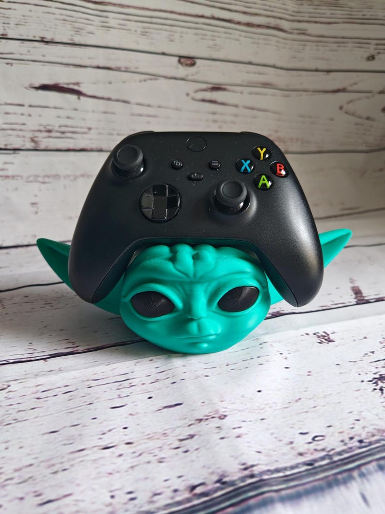 Op de afbeelding: Een zwarte Xbox-controller wordt in een groene Yoda-standaard gehouden. De controller heeft de standaard Xbox-knoppen: A, B, X, Y. De standaard is gemaakt van plastic en heeft een groen, buitenaards gezicht.