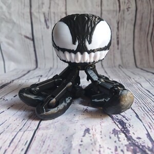 May include: A black and white 3D printed figurine of Venom, a fictional character from Marvel Comics. The figurine has a round head with a white face and black eyes, and a body with six articulated legs.