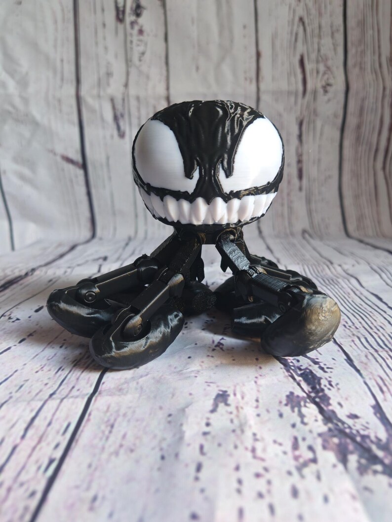 May include: A black and white 3D printed figurine of Venom, a fictional character from Marvel Comics. The figurine has a round head with a white face and black eyes, and a body with six articulated legs.