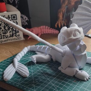 May include: A white 3D printed model of a dragon with wings outstretched. The dragon is posed on its belly with its head raised. The model is printed in multiple pieces and is ready to be assembled.