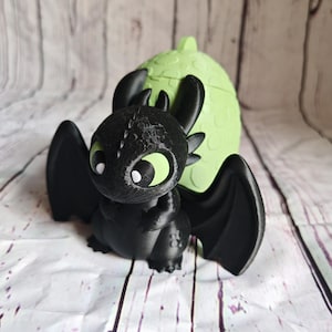 K&ouml;nnte beinhalten: Schwarze Drachenfigur mit gro&szlig;en Fl&uuml;geln und gr&uuml;nen Augen, vor einem hellgr&uuml;nen, eif&ouml;rmigen Beh&auml;lter. Der Drache hat detaillierte Texturen und einen freundlichen Ausdruck. Der Beh&auml;lter hat eine strukturierte Oberfl&auml;che.