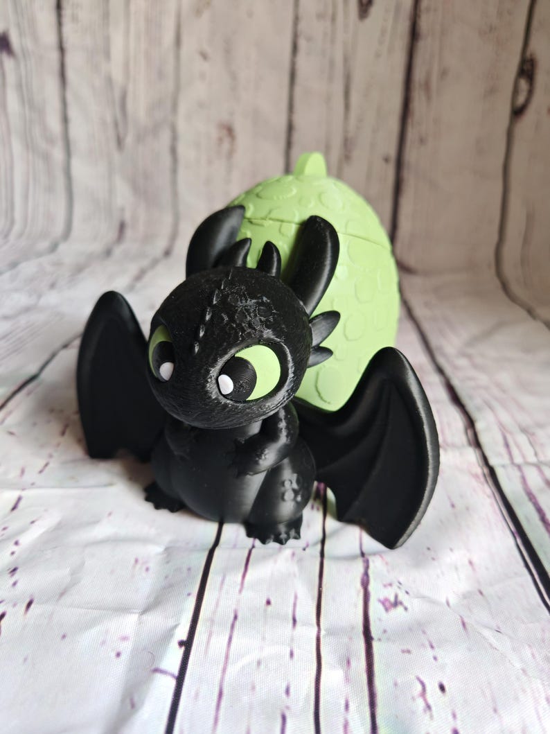 K&ouml;nnte beinhalten: Schwarze Drachenfigur mit gro&szlig;en Fl&uuml;geln und gr&uuml;nen Augen, vor einem hellgr&uuml;nen, eif&ouml;rmigen Beh&auml;lter. Der Drache hat detaillierte Texturen und einen freundlichen Ausdruck. Der Beh&auml;lter hat eine strukturierte Oberfl&auml;che.
