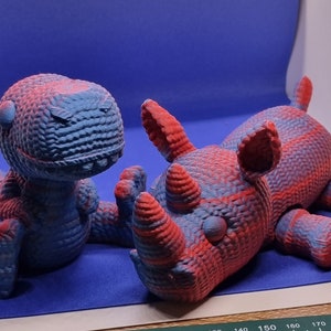 May include: Two 3D printed animal figurines, a blue and red dinosaur and a red and blue rhinoceros, are lying on a blue surface.