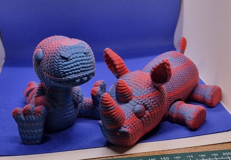 May include: Two 3D printed animal figurines, a blue and red dinosaur and a red and blue rhinoceros, are lying on a blue surface.