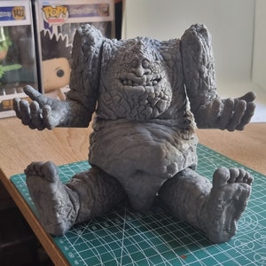 May include: A gray, textured, 3D printed figure of a creature with a large, toothy grin. The figure is sitting on a green cutting mat with white markings.