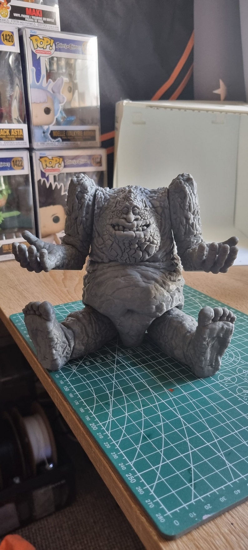 May include: A gray, textured, 3D printed figure of a creature with a large, toothy grin. The figure is sitting on a green cutting mat with white markings.