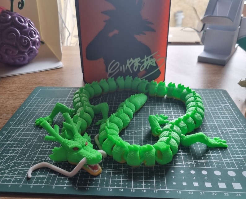 Op de afbeelding: Een groene drakenfiguur met een witte onderkant, gemaakt van gesegmenteerde stukken, die lijkt op een populair anime-personage. De draak is in een opgerolde positie op een groene snijmat geplaatst.