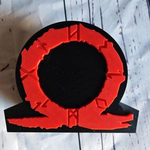 Peut inclure: Une figurine en 3D imprim&eacute;e en noir et rouge du logo God of War. Le logo est un cercle avec un anneau rouge autour et un symbole rouge en bas.