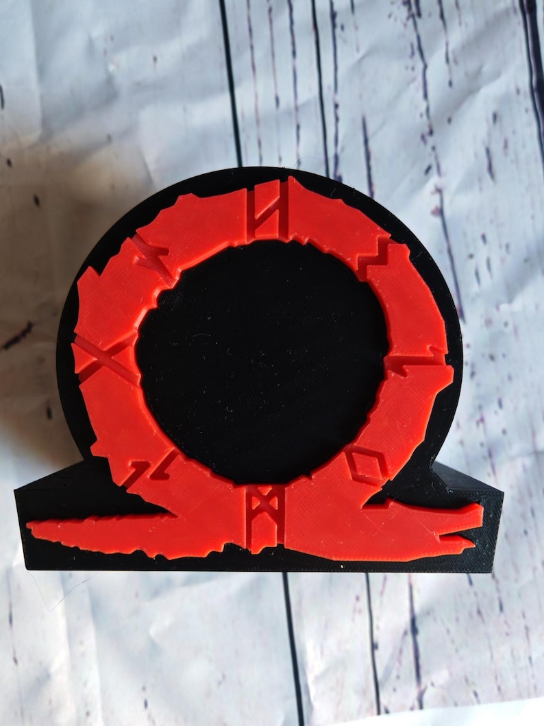 Peut inclure: Une figurine en 3D imprim&eacute;e en noir et rouge du logo God of War. Le logo est un cercle avec un anneau rouge autour et un symbole rouge en bas.