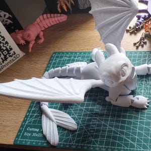 May include: A white 3D printed model of a dragon with wings spread out. The dragon is lying on a green cutting mat. The model is made up of multiple pieces that have been assembled together.