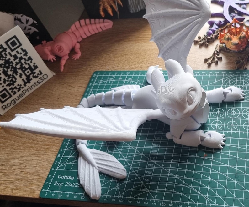 May include: A white 3D printed model of a dragon with wings spread out. The dragon is lying on a green cutting mat. The model is made up of multiple pieces that have been assembled together.