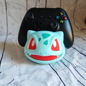 Puede incluir: Una figura de personaje Pokemon azul y roja con un mando Xbox negro encima. La figura es un Bulbasaur, un Pokemon de tipo planta.