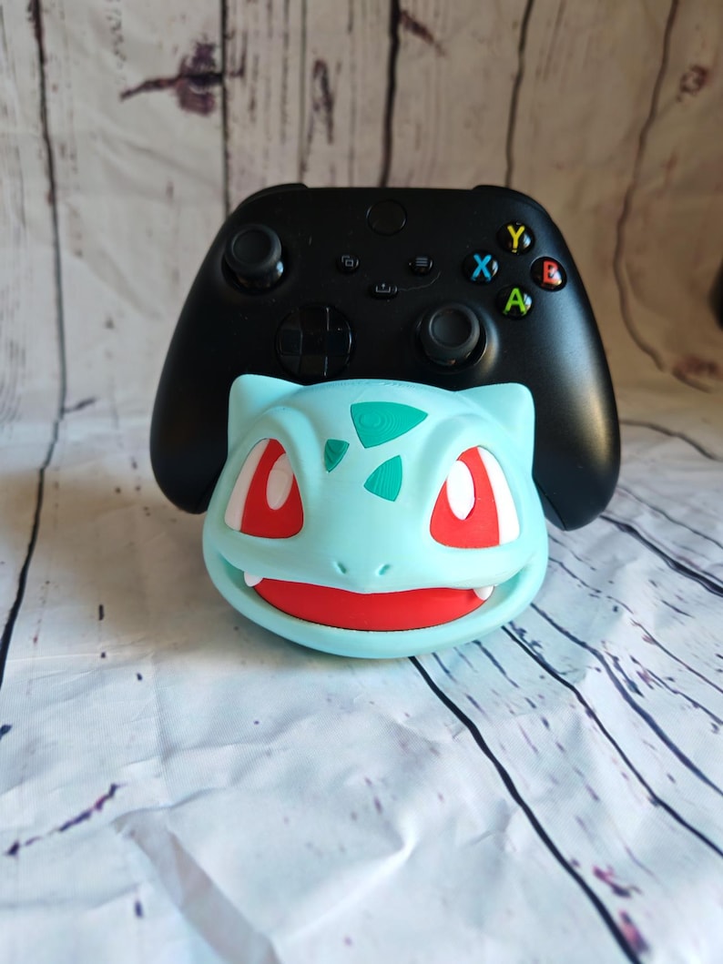 Puede incluir: Una figura de personaje Pokemon azul y roja con un mando Xbox negro encima. La figura es un Bulbasaur, un Pokemon de tipo planta.