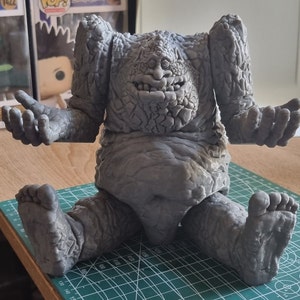 May include: A gray, stone-like figure with a large head and a wide, toothy grin. The figure is sitting on a green mat with a grid pattern.