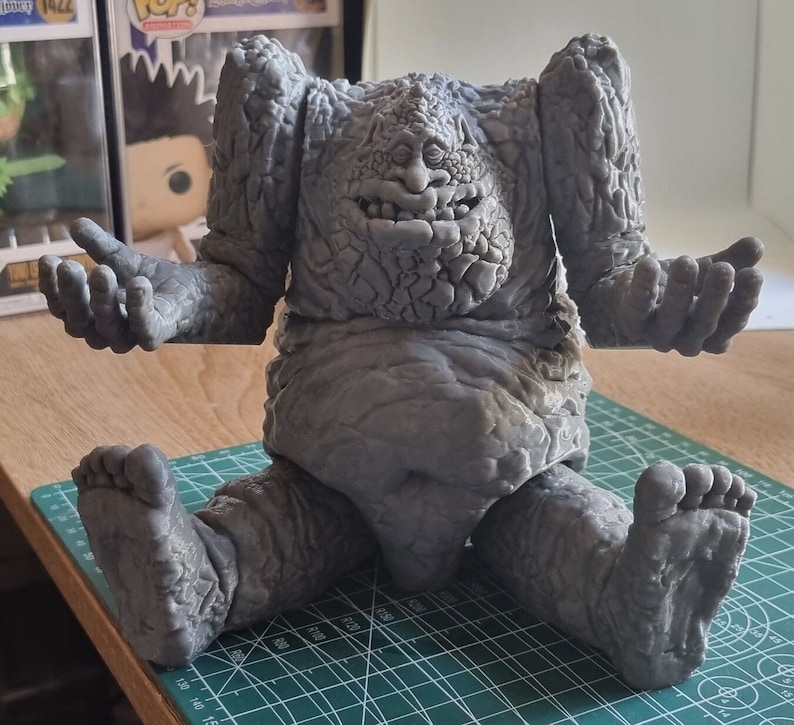 May include: A gray, stone-like figure with a large head and a wide, toothy grin. The figure is sitting on a green mat with a grid pattern.