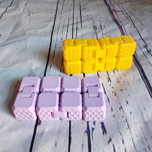 Peut inclure: Deux jouets anti-stress, l'un violet et l'autre jaune, compos&eacute;s de carr&eacute;s imbriqu&eacute;s avec un motif en nid d'abeille.