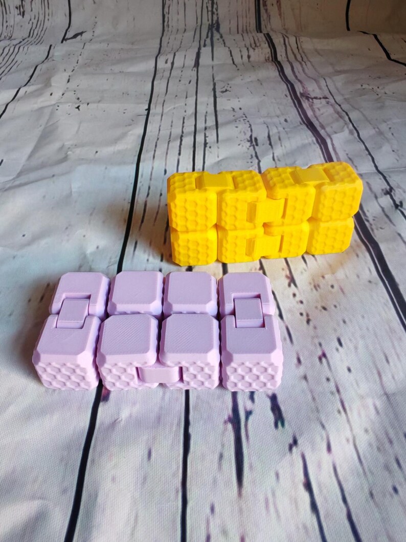 Peut inclure: Deux jouets anti-stress, l'un violet et l'autre jaune, compos&eacute;s de carr&eacute;s imbriqu&eacute;s avec un motif en nid d'abeille.
