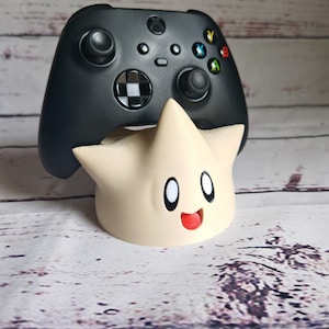May include: A black video game controller sits on top of a white, star-shaped stand with a cartoon face. The stand has a red mouth and black eyes.