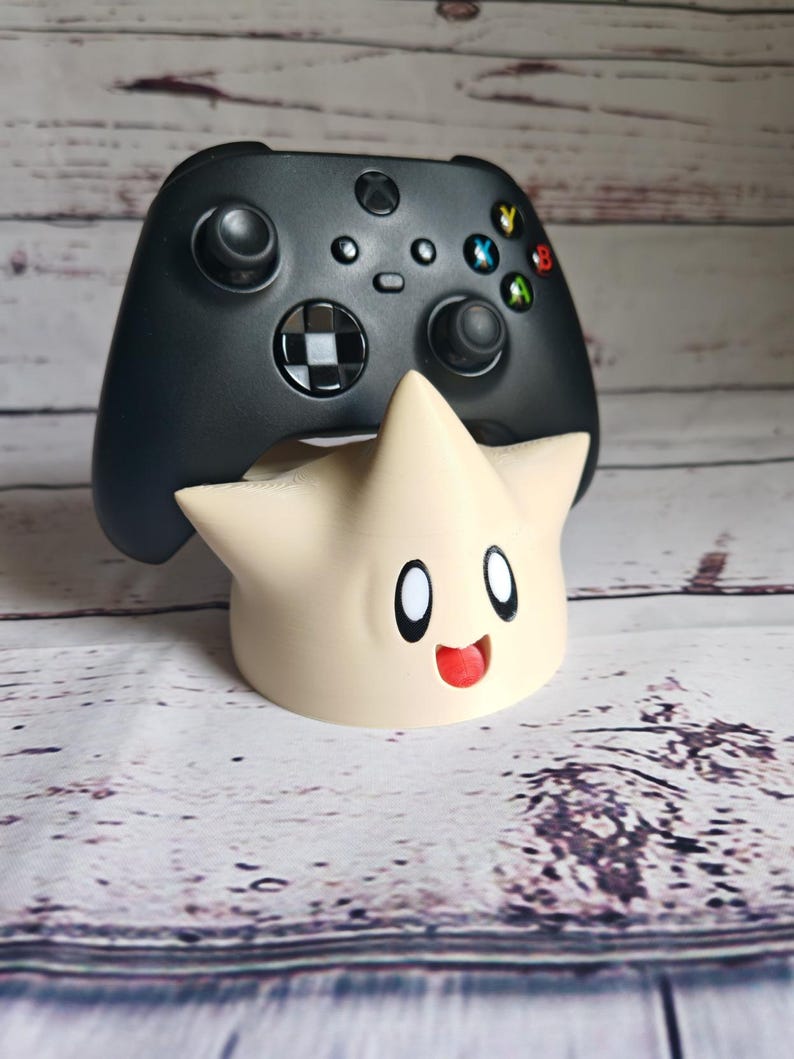 May include: A black video game controller sits on top of a white, star-shaped stand with a cartoon face. The stand has a red mouth and black eyes.