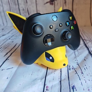 May include: A black Xbox controller is held in a yellow and black 3D printed stand shaped like a cartoon character. The stand has a blue eye and a yellow nose.